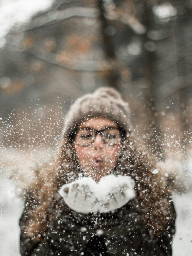 A woman in winter clothing playfully blowing snowflakes outdoors, capturing the joy of a snowy day.