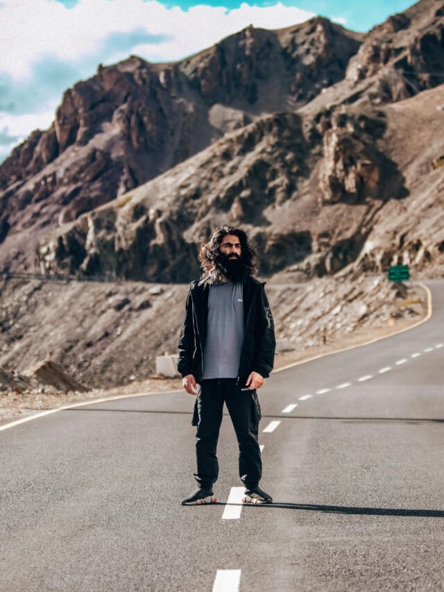 A person stands on a winding road in Kargil with scenic rocky mountains in the background.