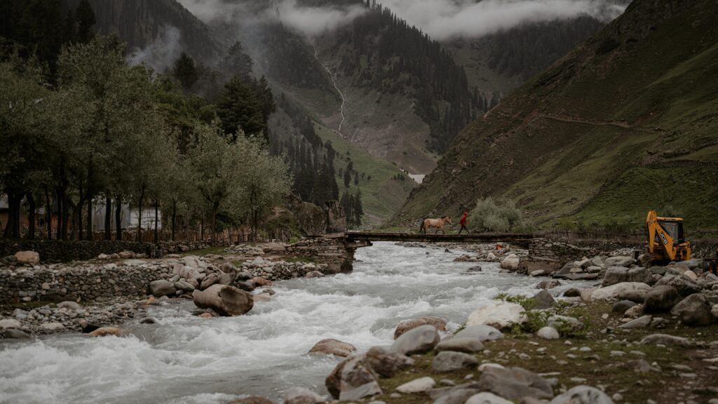 Bridge in Gulmarg A tranquil scene of a person leading a horse over a wooden bridge in Kashmir's lush, mountainous landscape. Gulmarg: Complete Travel Guide.